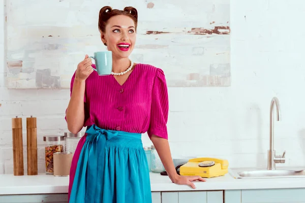 微笑别针女孩在深红色礼服和蓝色围裙站立在厨房桌与咖啡杯 — 图库照片