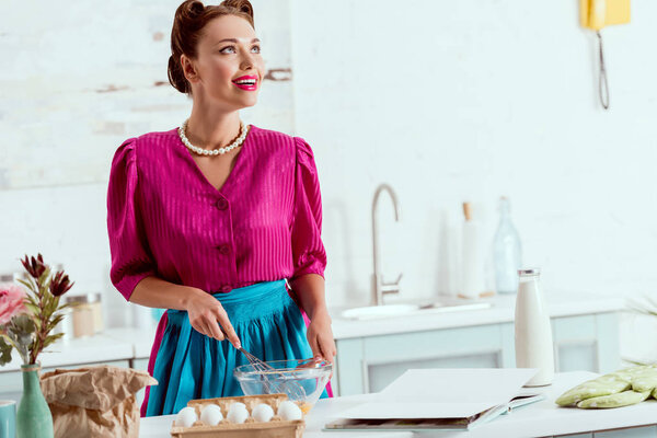 Улыбающаяся девушка в малиновом платье и синем фартуке смешивая ингредиенты для приготовления теста
 