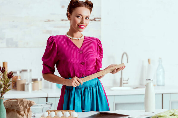 Элегантно одетый пин-ап девушка держит деревянную скалку и смотрит в камеру
