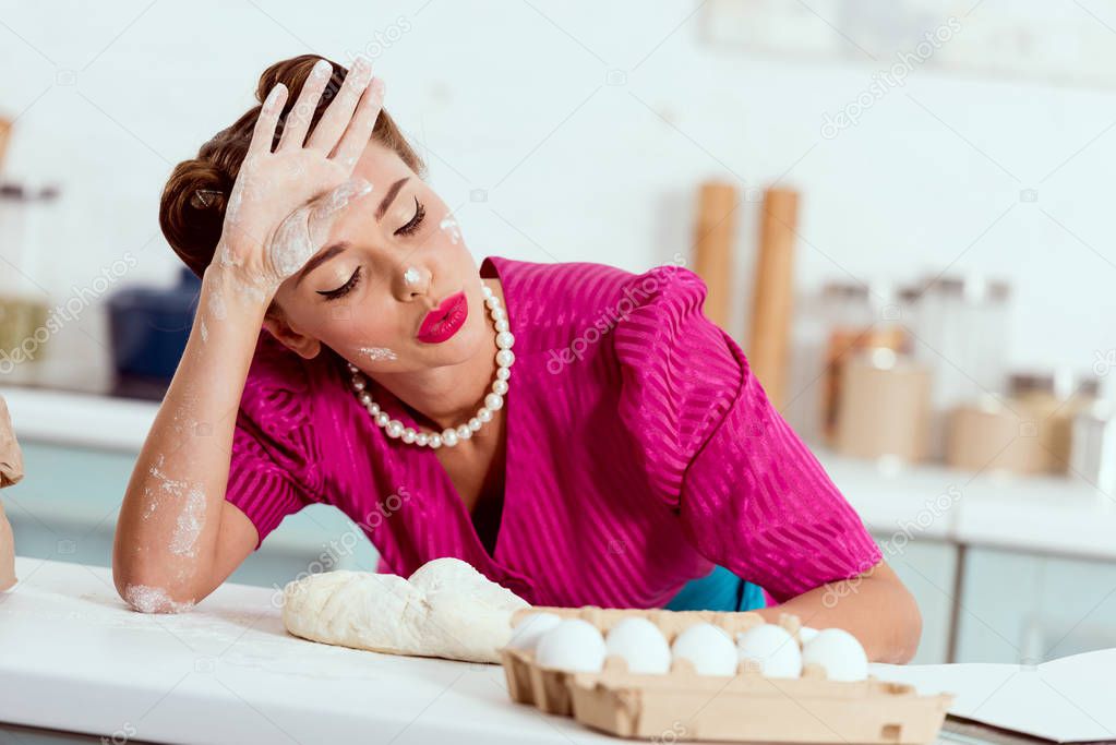 exhausted pin up girl with flour traces on hands and face leaning on kitchen table