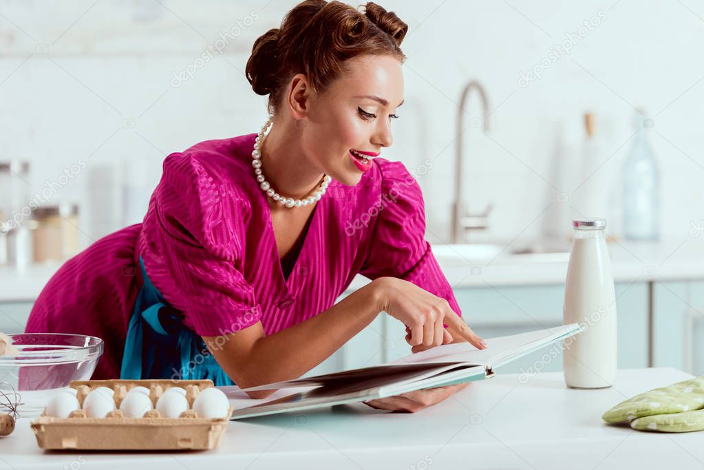 Elegant smiling pin up girl reading recipes book while leaning on kitchen table 