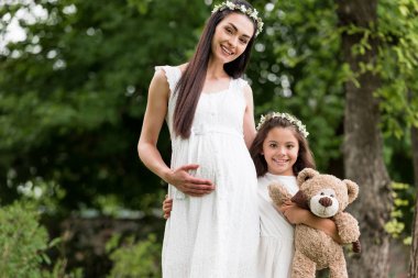 mutlu hamile anne ve sevimli kızı teddy ile birlikte duran ve kamera Parkta gülüp ayı