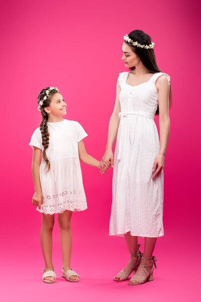 beautiful happy mother and daughter in white dresses and floral wreaths holding hands and smiling each other on pink
