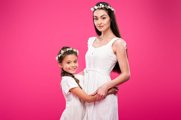 beautiful happy mother and daughter in white dresses and floral wreaths hugging and smiling at camera isolated on pink