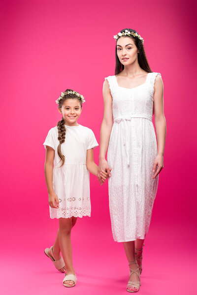 beautiful happy mother and daughter in white dresses and floral wreaths holding hands and smiling at camera on pink
