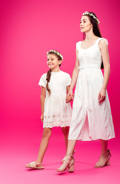 beautiful happy mother and daughter in white dresses and floral wreaths holding hands and walking on pink