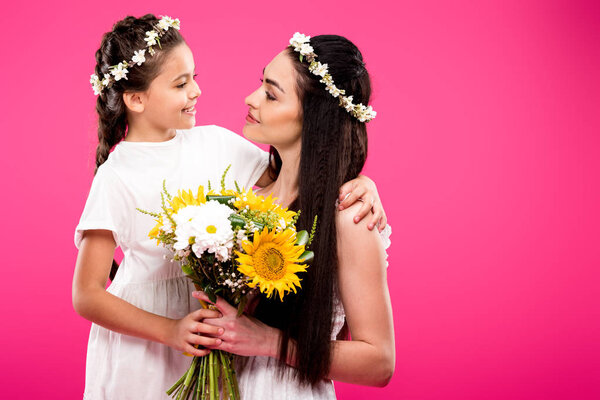 beautiful happy mother and daughter in white dresses holding flower bouquet and smiling each other isolated on pink