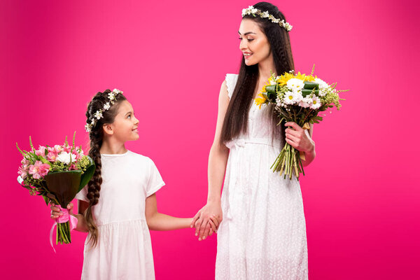 beautiful happy mother and daughter in white dresses and floral wreaths holding bouquets and smiling each other isolated on pink