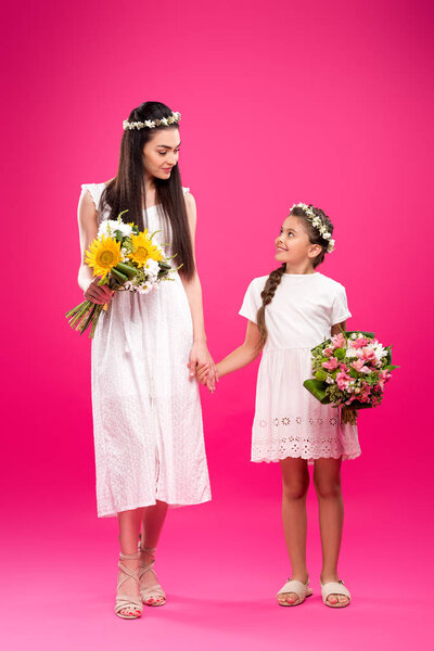 full length view of beautiful mother and daughter in white dresses holding bouquets and smiling each other on pink