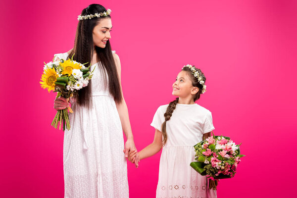 beautiful mother and daughter in white dresses holding bouquets and smiling each other isolated on pink