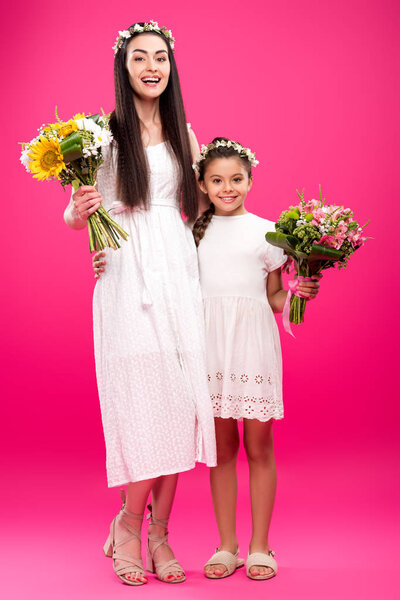 happy mother and daughter in white dresses and floral wreaths holding beautiful bouquets on pink