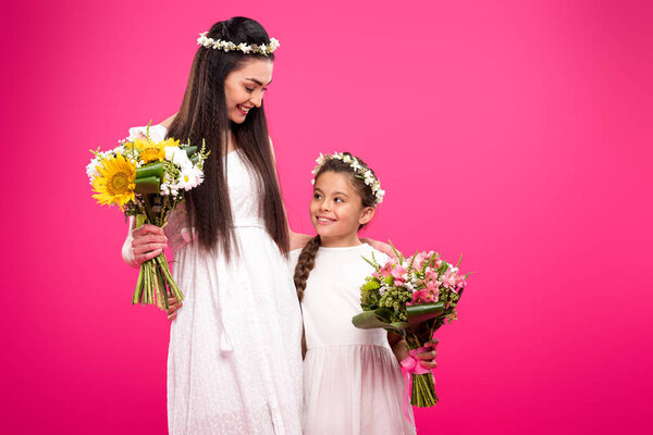 beautiful happy mother and daughter in white dresses and floral wreaths holding bouquets isolated on pink