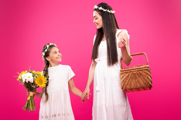 beautiful happy mother and daughter in white dresses holding bouquet and picnic basket isolated on pink 