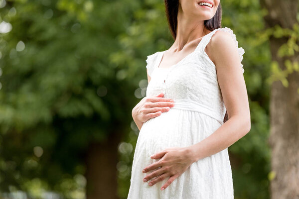 cropped shot of smiling pregnant woman in white dress touching belly in park 