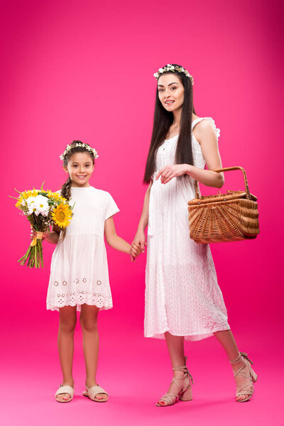 beautiful happy mother and daughter in white dresses holding bouquet and picnic basket on pink