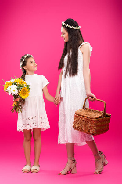 beautiful happy mother and daughter in white dresses holding bouquet and picnic basket on pink 