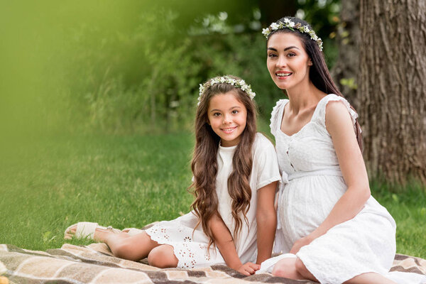 happy pregnant mother and cute daughter in white dresses and wreaths sitting together on plaid and smiling at camera in park