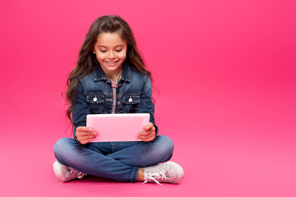 adorable happy child in denim clothes sitting and using digital tablet on pink