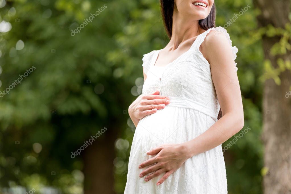 cropped shot of smiling pregnant woman in white dress touching belly in park 