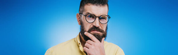 suspicious bearded man in eyeglasses looking at camera isolated on blue
