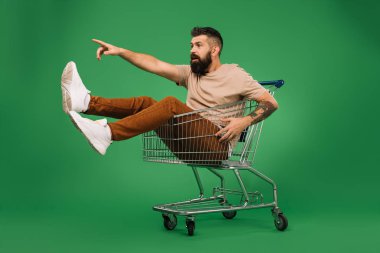 excited handsome man showing something while sitting in shopping cart isolated on green clipart