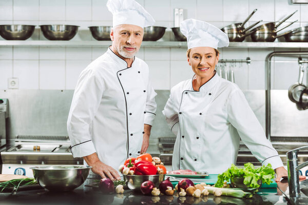 female and male chefs in uniform and hats looking at camera during cooking in restaurant kitchen
