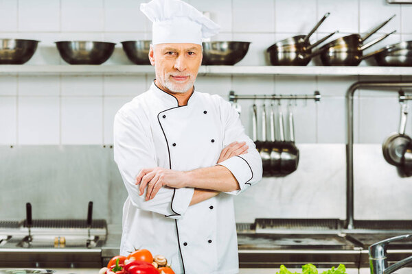 handsome male chef in uniform with arms crossed looking at camera in restaurant kitchen