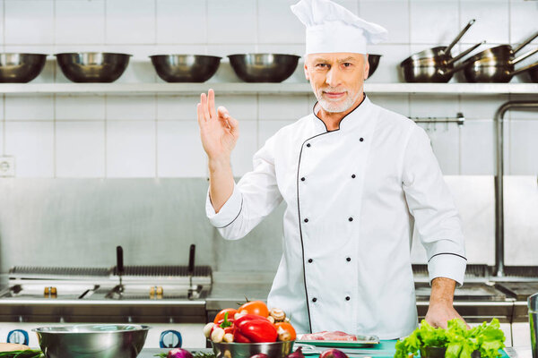 handsome smiling male chef in uniform looking at camera and showing ok sign in restaurant kitchen