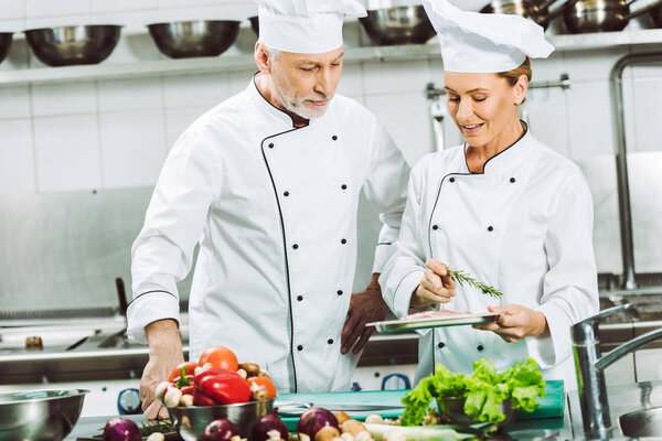 female and male chefs in double-breasted jackets and hats decorating dish with rosemary while cooking in restaurant kitchen