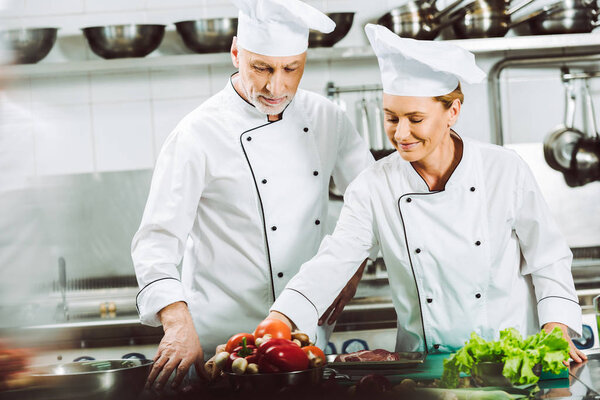 selective focus of female and male chefs in uniform and hats cooking in restaurant kitchen