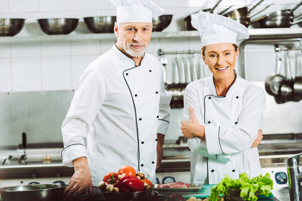 smiling female and male chefs in uniform and hats looking at camera while cooking in restaurant kitchen