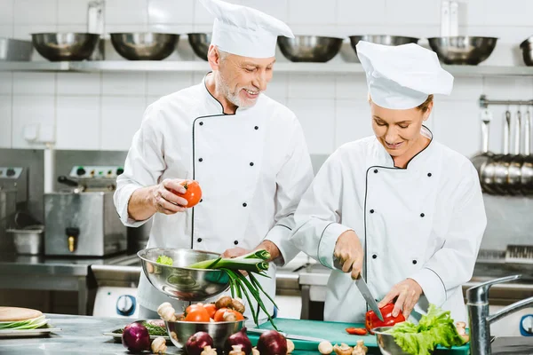 微笑的女性和男性厨师在双排扣夹克和帽子烹饪在餐厅厨房 — 图库照片