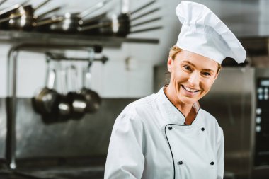 beautiful female chef in uniform and hat looking at camera in restaurant kitchen with copy space clipart