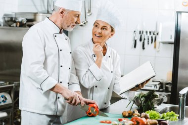 Restoran mutfağında yemek sırasında yemek tarifi kitabı kullanarak üniformalı kadın ve erkek şefler