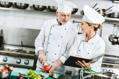 Restoran mutfağında yemek sırasında yemek tarifi kitabı kullanarak üniformalı kadın ve erkek şefler