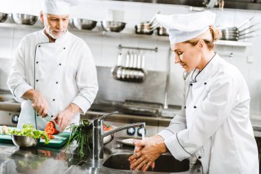 selective focus of male and female chefs in double-breasted jackets cooking in restaurant kitchen clipart