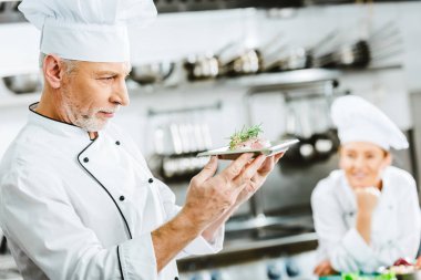 düşünceli erkek aşçı ile arka plan üzerinde meslektaşım plaka üzerinde et yemek Restoran mutfağında tutan üniformalı