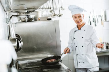 beautiful smiling female chef in uniform looking at camera while cooking meat in restaurant kitchen clipart