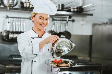beautiful smiling female chef in uniform holding dome from serving tray with meat dish in restaurant kitchen clipart