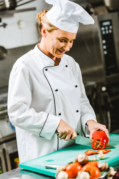 beautiful smiling female chef in uniform cutting pepper in restaurant kitchen