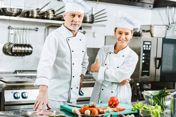 female and male chefs in uniform looking at camera during cooking in restaurant kitchen