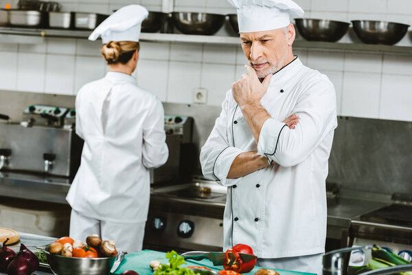 pensive male chef in double-breasted jacket during cooking in restaurant kitchen with female colleague on background