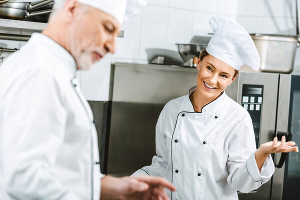 selective focus of female and male chef in uniforms having conversation in restaurant kitchen