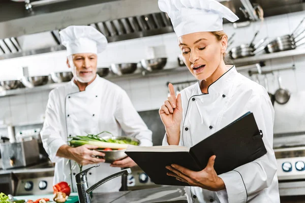 Female Chef Uniform Holding Recipe Book Doing Idea Gesture While — Stock Photo, Image