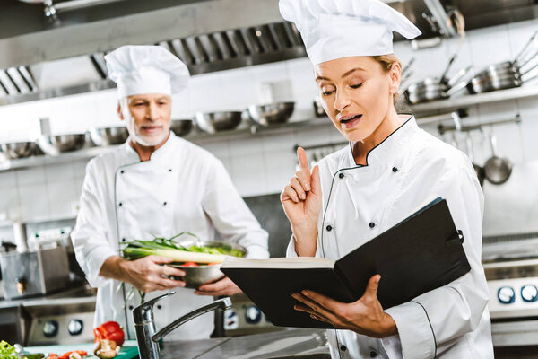female chef in uniform holding recipe book and doing idea gesture while colleague cooking on background in restaurant kitchen