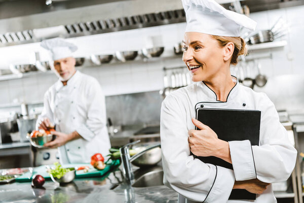 beautiful female chef in uniform holding recipe book in restaurant kitchen with colleague on background
