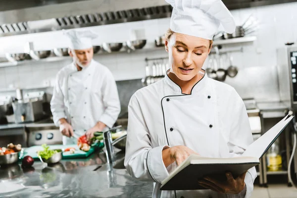 Žena Kuchař Uniformě Čtení Knihy Recept Zatímco Kolega Vaření Pozadí — Stock fotografie