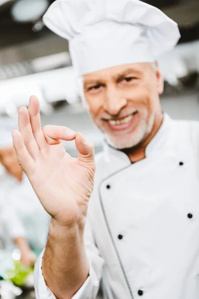 英俊的微笑的男性厨师在制服看着相机 并显示确定标志在餐厅厨房 — 图库照片