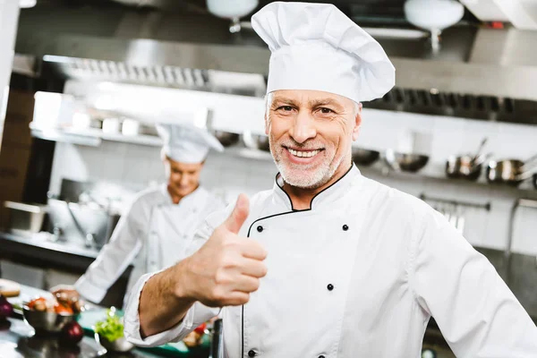 レストランのキッチンでハンサムな制服を着た男性シェフの笑みを浮かべてカメラを見て 親指を現してサインします — ストック写真