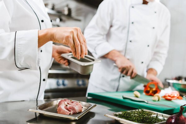 cropped view of female chef seasoning meat while man cooking on background in restaurant kitchen